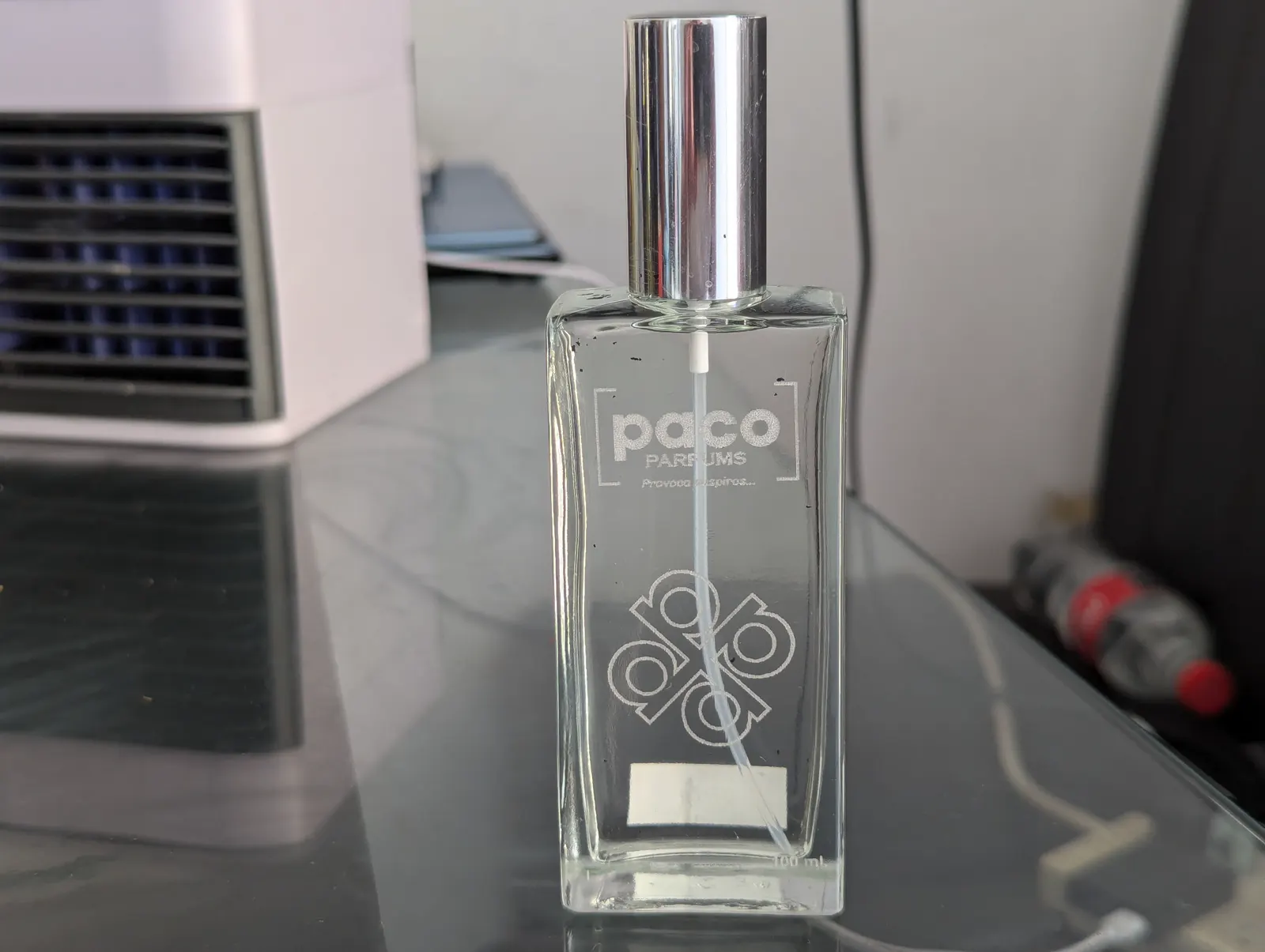 Perfume fotografiado con celular sobre un escritorio, con un mini clima y un refresco al fondo.